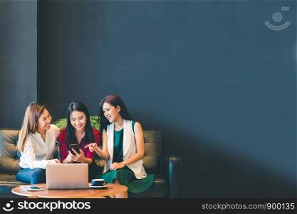 Three beautiful Asian girls using smartphone and laptop, chatting on sofa together at cafe with copy space, modern lifestyle with gadget technology or working woman on casual business concept