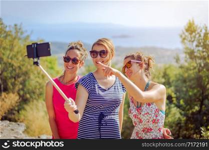 Three beautiful girls female travelers in summer dresses taking selfies with smartphone using selfie stick friends friendship outdoors on the mountain above the sea in autumn