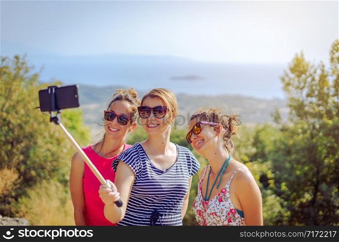 Three beautiful girls female travelers in summer dresses taking selfies with smartphone using selfie stick friends friendship outdoors on the mountain above the sea in autumn