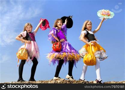 Three Beautiful Smiling Little Girls with Long Hair in the Colorful Witch Costume Greeting with Trick or Treat Bags at the Blue Sky.