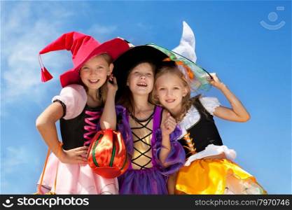 Three Beautiful Smiling Little Girls with Long Hair in the Witch Costume Looking at Camera with Trick or Treat Bags at the Blue Sky.&#xA;