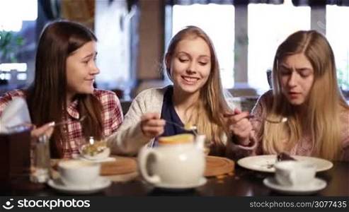 Three beautiful teenage females having great time together in cafe. Smiling attractive young women chatting about latest gossips in coffee shop and laughing while eating desserts in coffee shop after lessons.