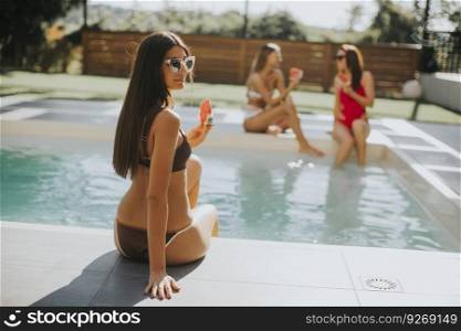 Three beautiful young woman with sun glasses sitting on poolside and eating watermelon at a resort swimming pool on a sunny day