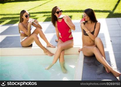 Three beautiful young woman with sun glasses sitting on poolside and eating watermelon at a resort swimming pool on a sunny day
