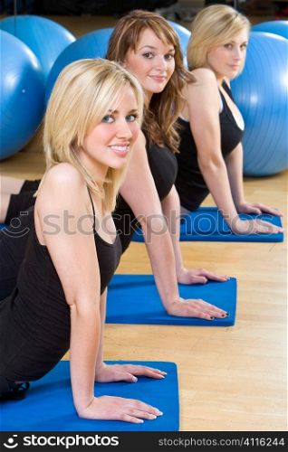 Three beautiful young women, two blond, one brunette, working out on on mats doing aerobic stretching at the gym. The focus is on the blond girl with blue eyes in the foreground.