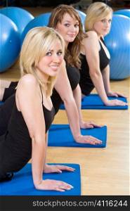 Three beautiful young women, two blond, one brunette, working out on on mats doing aerobic stretching at the gym. The focus is on the blond girl with blue eyes in the foreground.