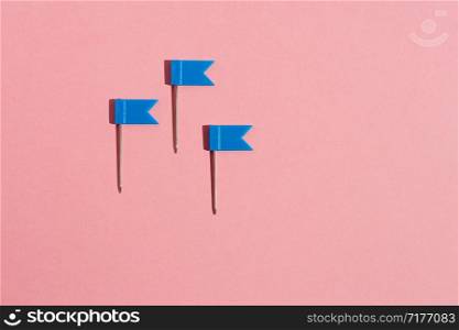 Three blue little flag pins on a pink background. View from above .. Three blue little flag pins on a pink background. View from above