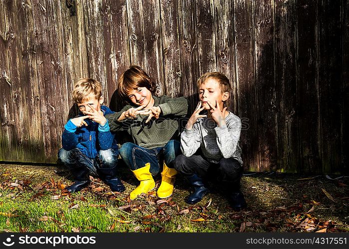 Three boys, outdoors, crouching beside fence, making hand gestures