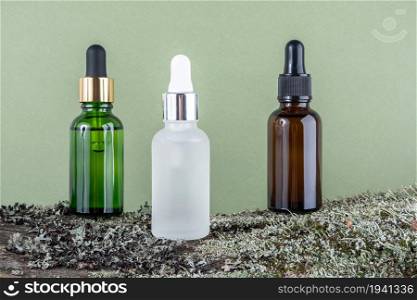Three brown, green, white glass bottles with serum, essential oil or other cosmetic product on tree bark covered with moss against green background. Natural Organic Spa Cosmetic Beauty concept.. Three brown, green, white glass bottles with serum, essential oil or other cosmetic product on tree bark covered with moss against green background. Natural Organic Spa Cosmetic Beauty concept
