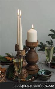Three burning candles on a table in wooden candlesticks, two glasses with champagne, spruce branches, green-brown ceramic cups.