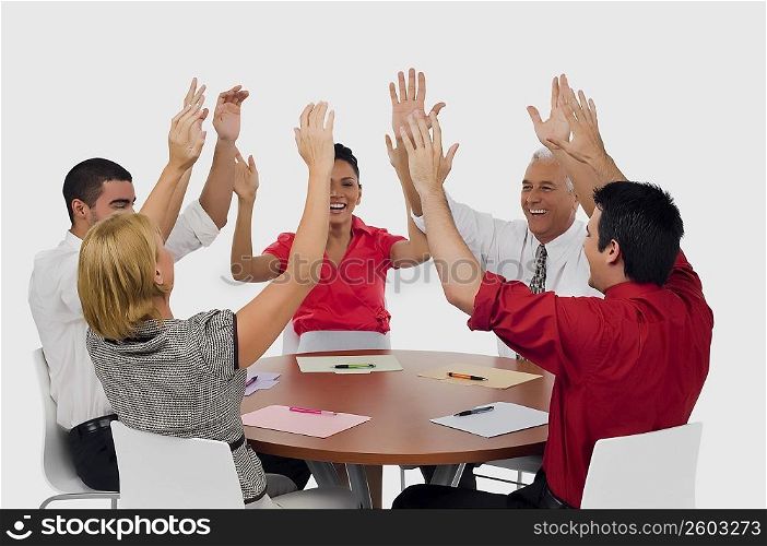 Three businessmen and two businesswomen smiling with their arms raised in a meeting