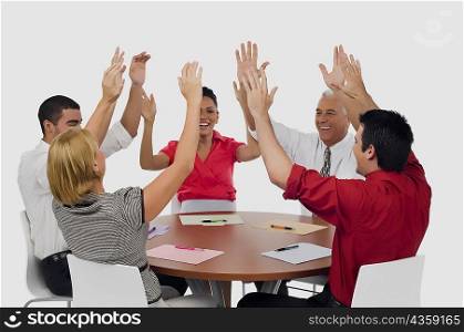 Three businessmen and two businesswomen smiling with their arms raised in a meeting
