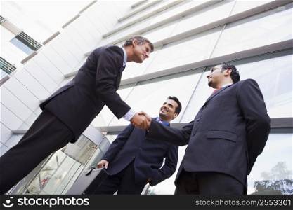 Three businessmen standing outdoors by building shaking hands and smiling (high key/selective focus)