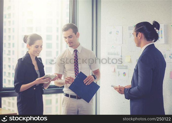 Three businesspeople standing in modern office looking file document and talking in meeting room.