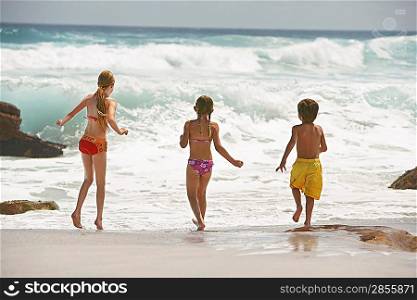 Three children (5-6 7-9 10-12) running on beach back view