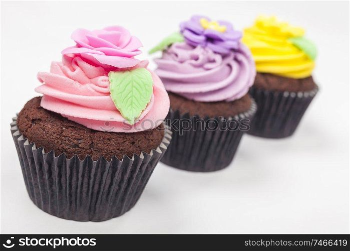 Three chocolate cup cakes with icing or frosting, pink, purple and yellow with green leaves, photographed on a white background