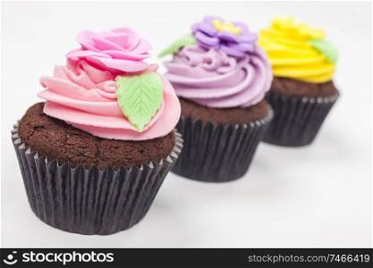 Three chocolate cup cakes with icing or frosting, pink, purple and yellow with green leaves, photographed on a white background