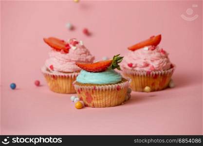 Three colorful cupcakes with strawberries on pink background with colorful candies