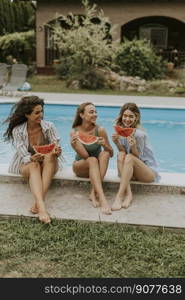 Three cute young women sitting on by the swimming pool and eating watermelon in the house backyard