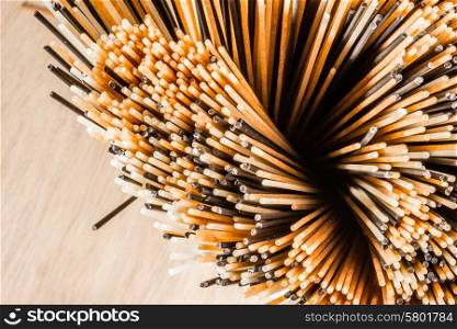 Three different colours of raw spaghetti sticks, all mixed together in brown, white, and black, are viewed from above as they stand up straight on a wooden table.
