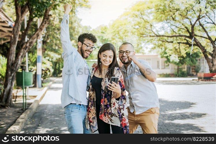 Three excited friends looking at the cell phone in the street. Three cheerful people looking at cell phone outdoors, Excited teenage friends looking at cell phone screen on the street