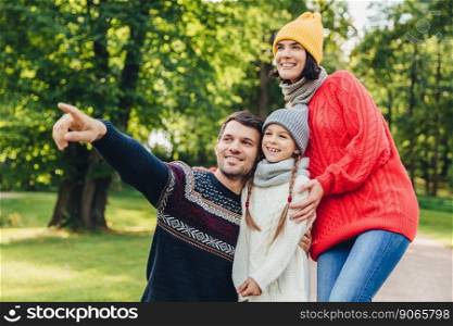 Three family members spend time together, look at beautiful lake in park, indicate with fingers, being in good mood, smile pleasantly. Father, mother and daughter enjoy togetherness, calm atmosphere
