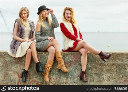 Three fashionable women sitting on concrete pier next to sea presenting pretty stylish outfits. Three fashionable woman on pier