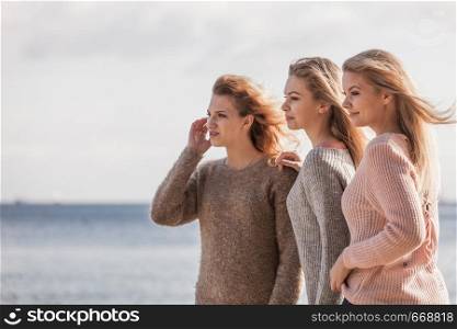 Three fashionable women wearing sweaters during warm autumnal weather spending their free time on sunny beach. Fashion models outdoor looking in distance. Three fashionable models outdoor