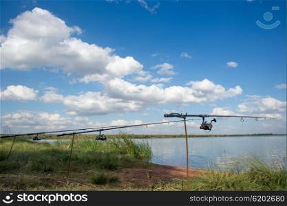 Three fishing rods are set up by the water of a lake or dam among green grass and in front of blue sky and white clouds.