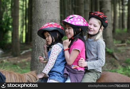 three friends: one caucasian blond and two asian (9 years +5 years) on horses in a forest