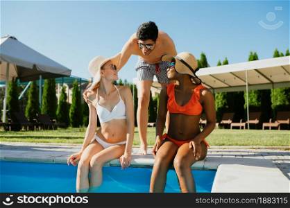 Three friends rest at the edge of the pool. Happy people having fun on summer vacations, holiday party at the poolside outdoors. One man and two women are sunbathing. Three friends rest at the edge of the pool