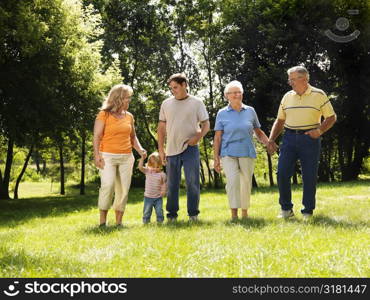 Three generation Caucasian family holding hands walking across grass in park smiling.