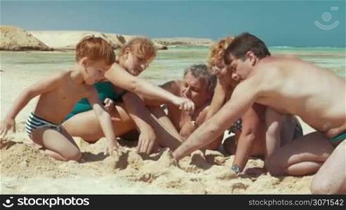 Three gengrations of one family are resting on the beach. They are playing with sand and pouring it.