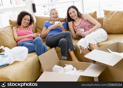 Three girl friends relaxing with coffee by boxes in new home smiling