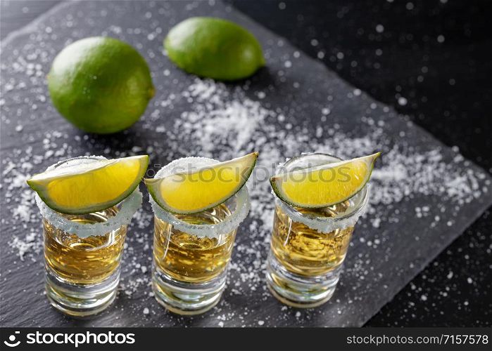 Three glasses of golden Mexican tequila and lime with salt on a dark background. Three glasses of golden Mexican tequila and lime with salt