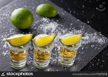 Three glasses of golden Mexican tequila and lime with salt on a dark background. Three glasses of golden Mexican tequila and lime with salt