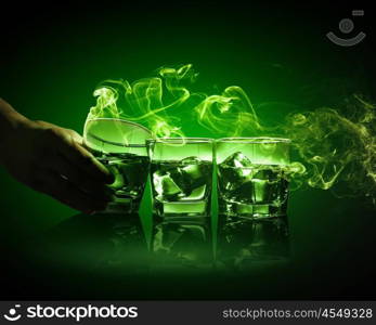 Three glasses of green absinth with ice cubes. Hand holding one of three glasses of green absinth with fume going out