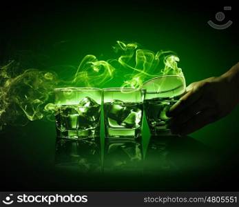 Three glasses of green absinth with ice cubes. Hand holding one of three glasses of green absinth with fume going out