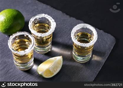 Three glasses of tequila with salt and pieces of lime on a black background. Alcoholic cocktail. Mexican traditional drink