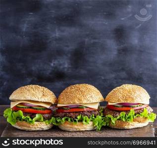 three hamburgers with vegetables on a brown wooden board, behind a black background, empty space