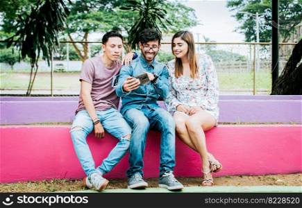 Three happy friends looking at the cell phone sitting in the park. Three cheerful people looking at cell phone sitting outside, happy teenage friends looking at phone screen outdoors