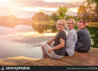 Three happy people sitting on the wooden pier during sunset