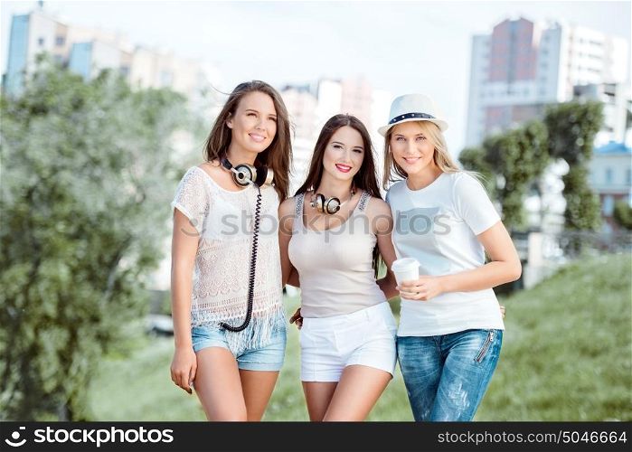 Three happy young women with vintage music headphones and a takeaway coffee cup, standing against urban city background and smiling.