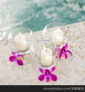Three lit candles in glass bowls with purple orchids next to pool.