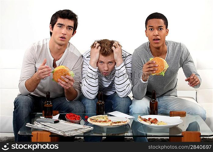Three male friends eating burgers and watching television