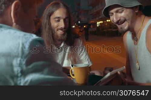 Three man friends are watching something in the white tablet computer speaking and smiling in summer cafe after sunset Thessaloniki, Greece