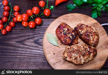 Three meat cutlets with spices on a wooden kitchen board with fresh vegetables, empty space on the left, top view