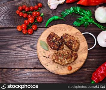 Three meat cutlets with spices on a wooden kitchen board with fresh vegetables, empty space on the left