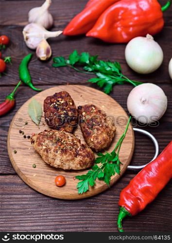 Three meat cutlets with spices on a wooden kitchen board with fresh vegetables, top view