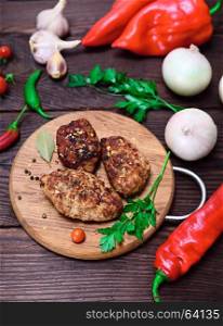 Three meat cutlets with spices on a wooden kitchen board with fresh vegetables, top view